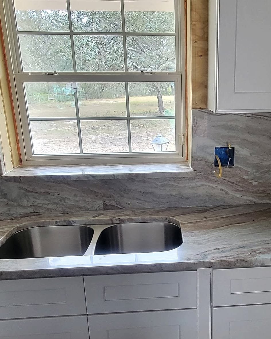 Marble countertop with a double undermount sink and full-height marble backsplash under a kitchen window, installed by Fuzion Stone Surfaces